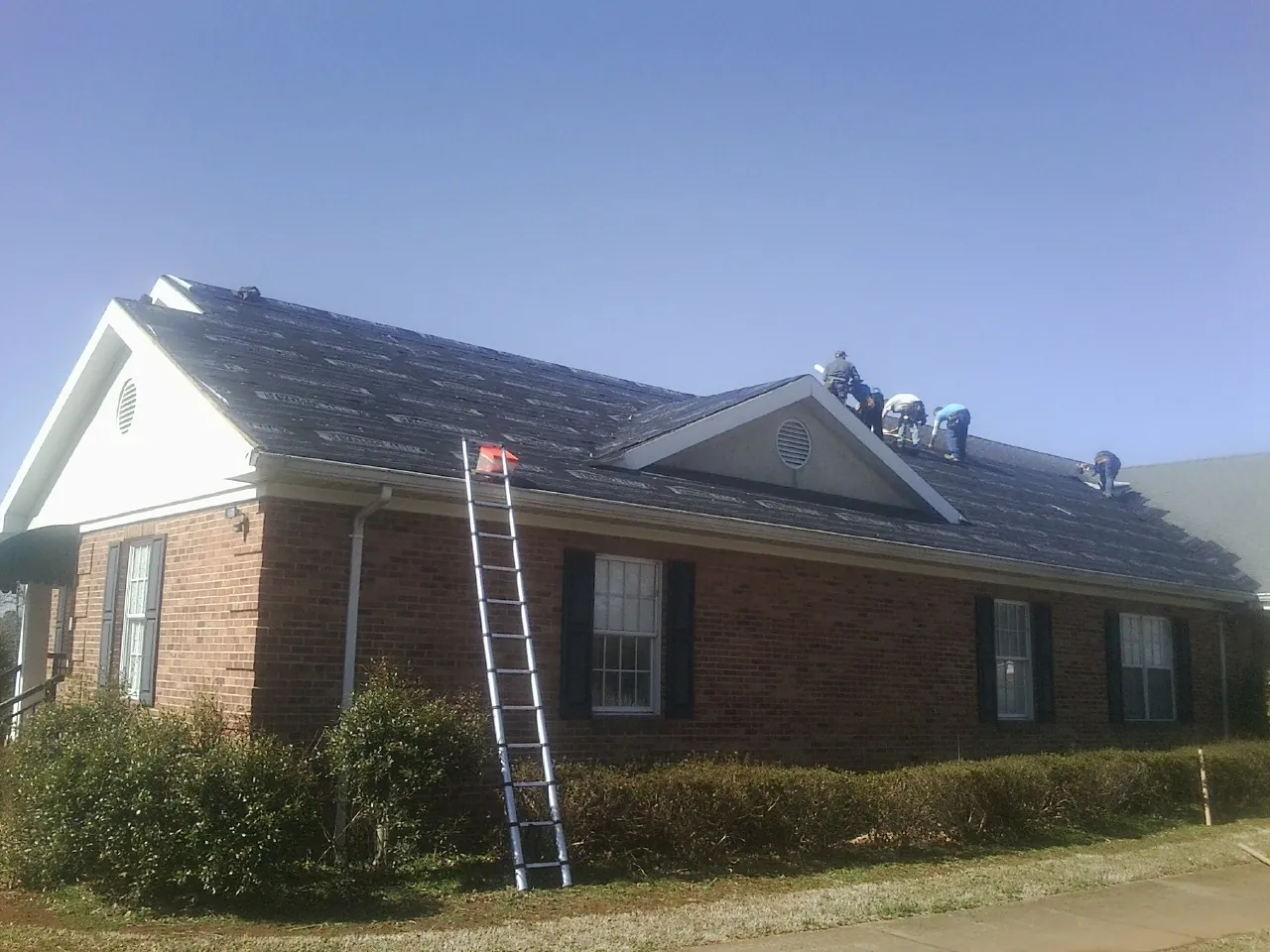 Crew tearing shingles off a brick ranch roof with a ladder set — Emergency Roof Repair work by our crew in New Prague