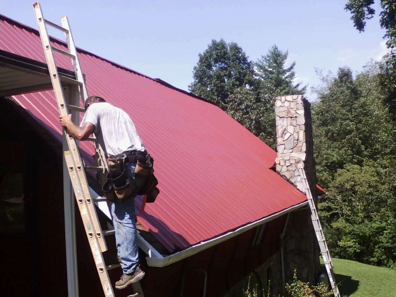 Roofer on a ladder beside a red metal roof and stone chimney — Soffit & Fascia Repair by Roofing Brothers in New Prague